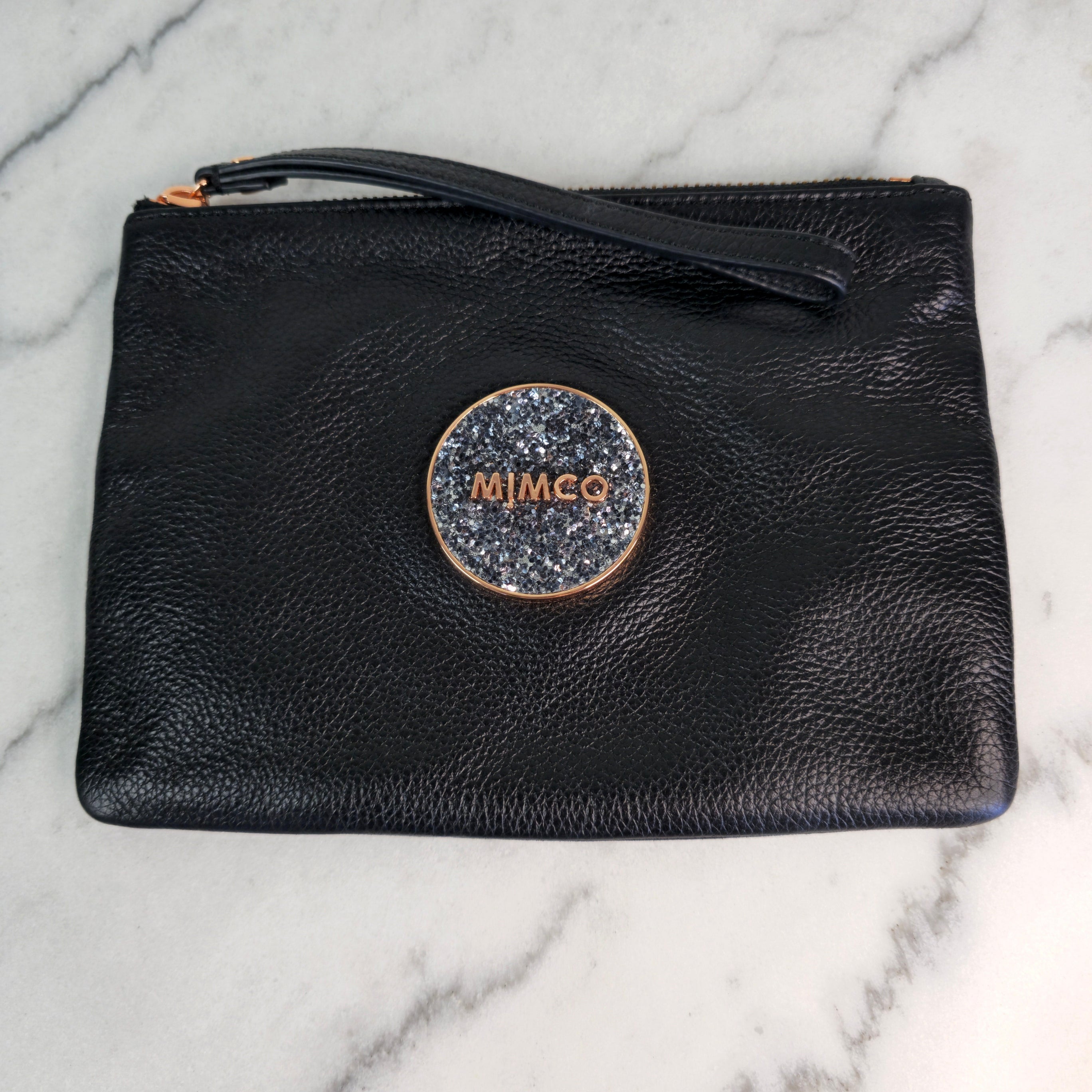 A black leather clutch pouch with rose gold accents, featuring the Mimco logo in a rose gold circular frame with a wrist strap on top.