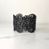 A black metal bracelet with an intricate lace-cut pattern, displayed on a marble surface.