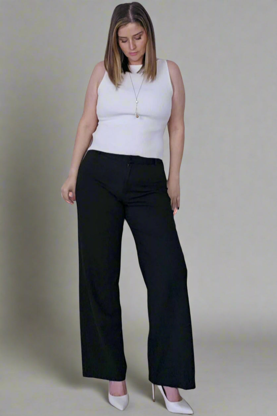 A woman standing against a plain background, wearing black dress pants and a white sleeveless top.