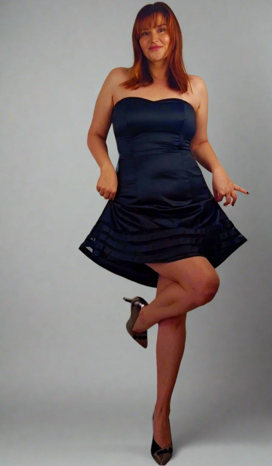 A woman posing in a black cocktail dress with a sweetheart neckline and sleeveless design.