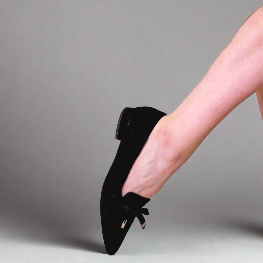 A close-up image of a black suede flat with the sole of the foot visible, against a background of tiled flooring.