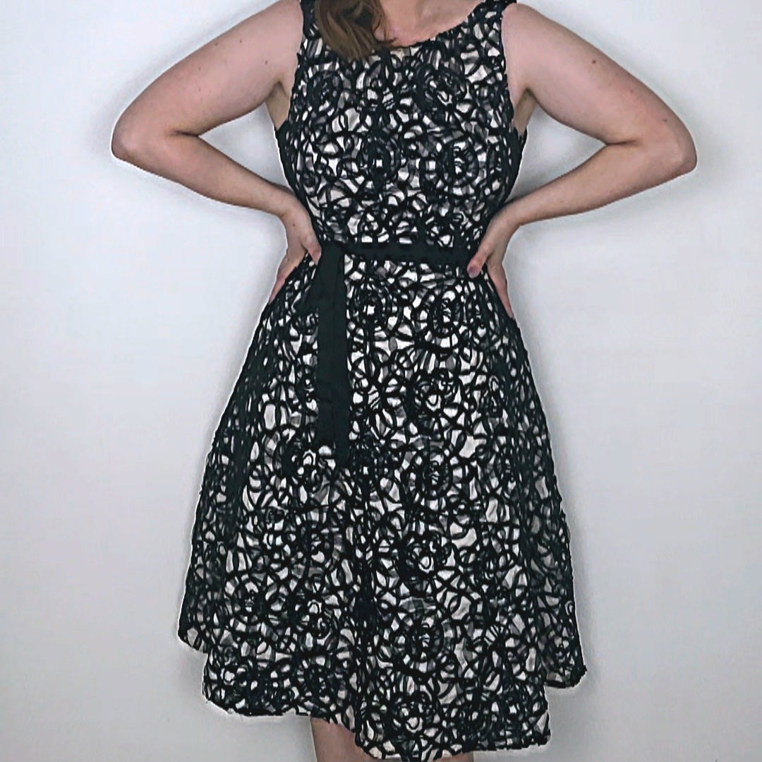 A model wearing a Phase Eight black and beige sleeveless dress with a round neckline, mesh floral pattern overlay, and a black ribbon-style belt at the waist.