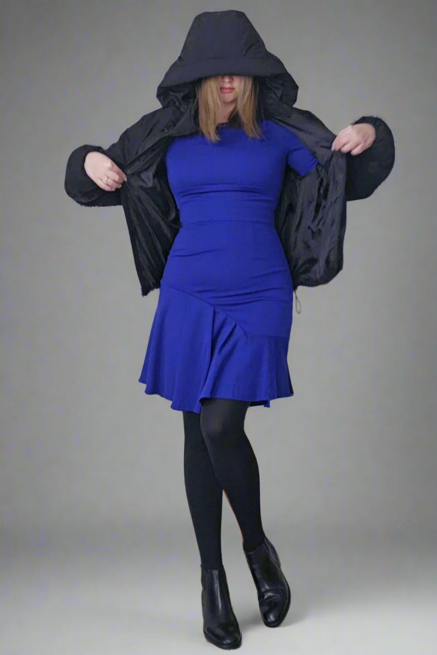 A person wearing a long sleeved, navy blue dress with an asymmetric hemline, standing in a room with a white wall and black jacket.
