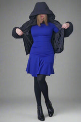 A person wearing a long sleeved, navy blue dress with an asymmetric hemline, standing in a room with a white wall and black jacket.