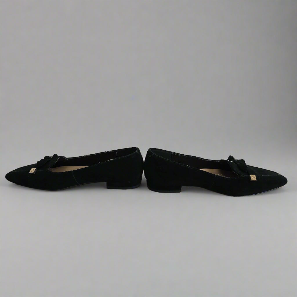 Black pointed-toe shoes with bow details leaning against a white wall.