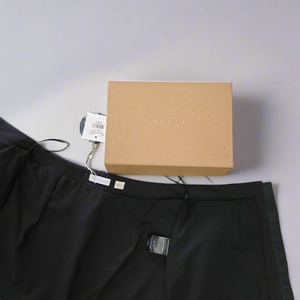 Black pants with a tag on a tiled floor next to a cardboard box.