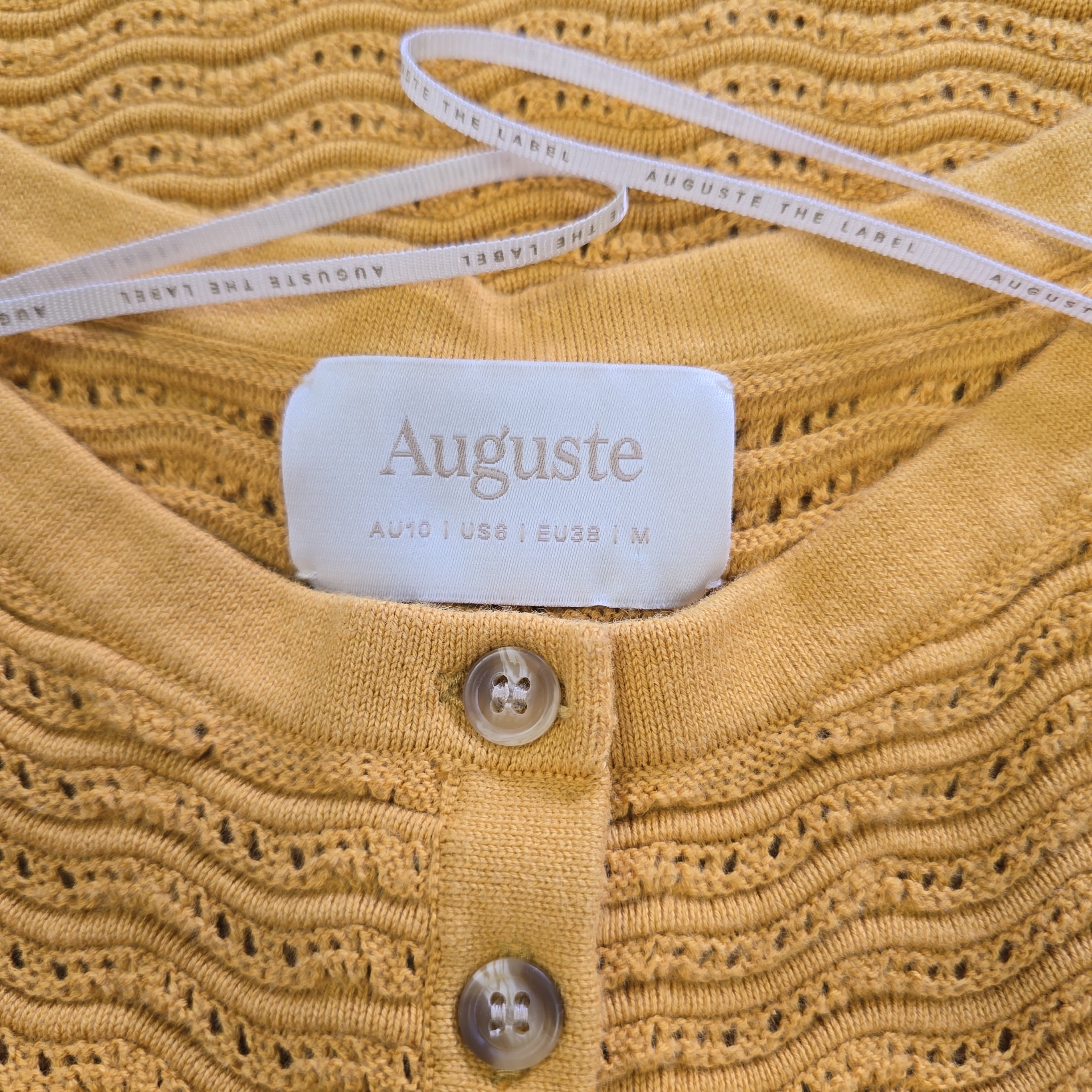 Close-up of a mustard yellow textured garment with 'Auguste' label and buttons.