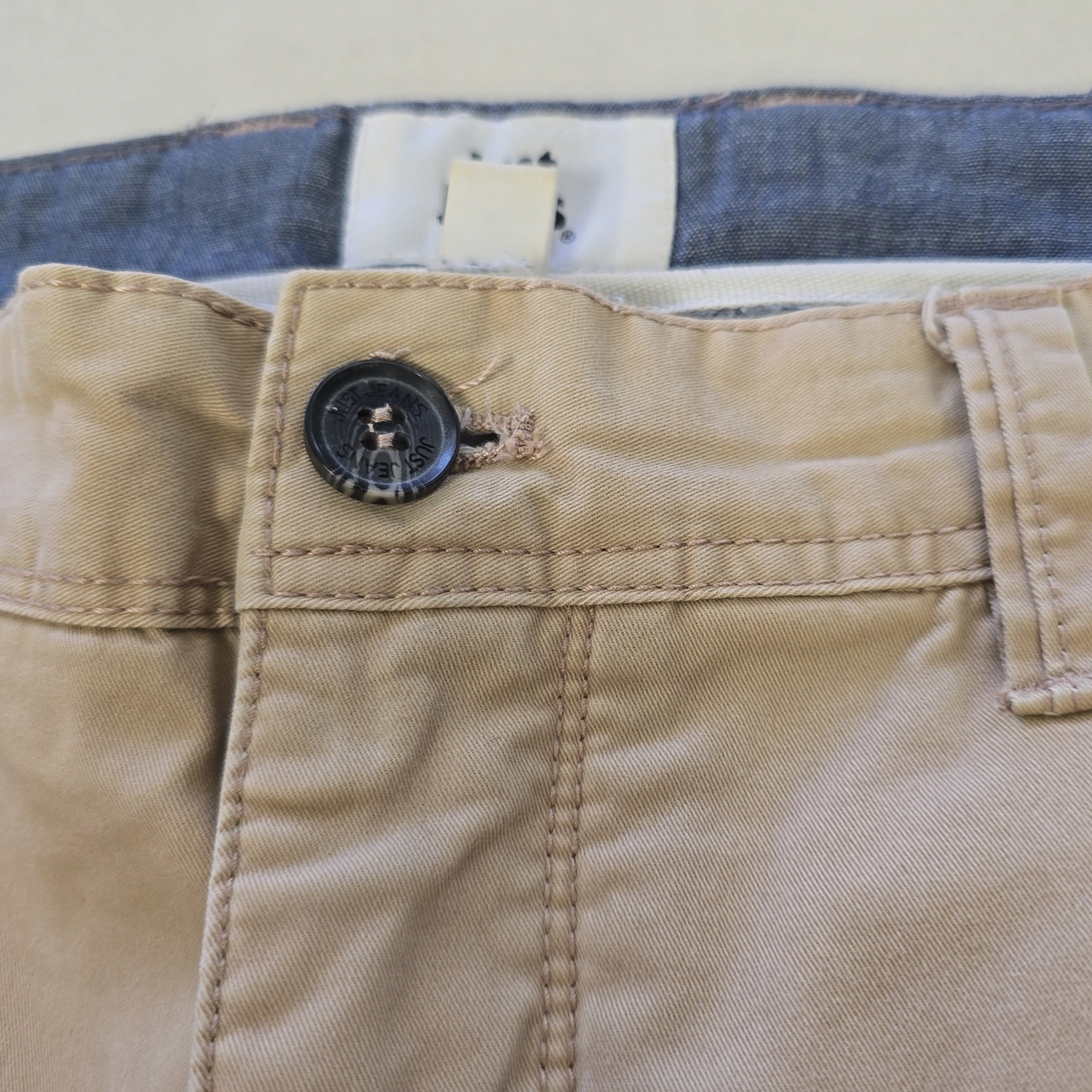 Close-up of beige pants with a button, with blue jeans partially visible in the background.