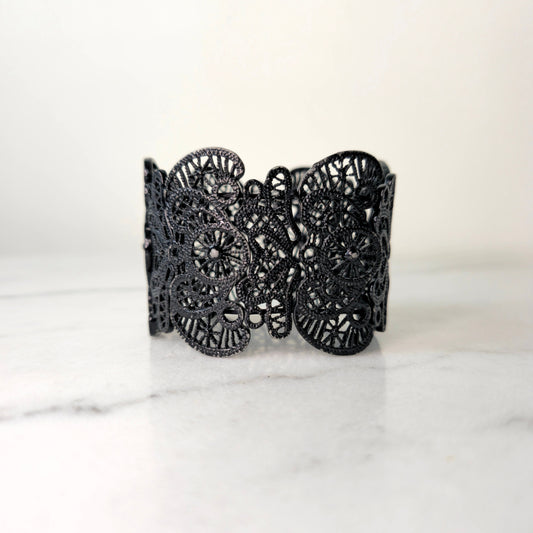 A black metal bracelet with an intricate lace-cut pattern, displayed on a marble surface. 2736